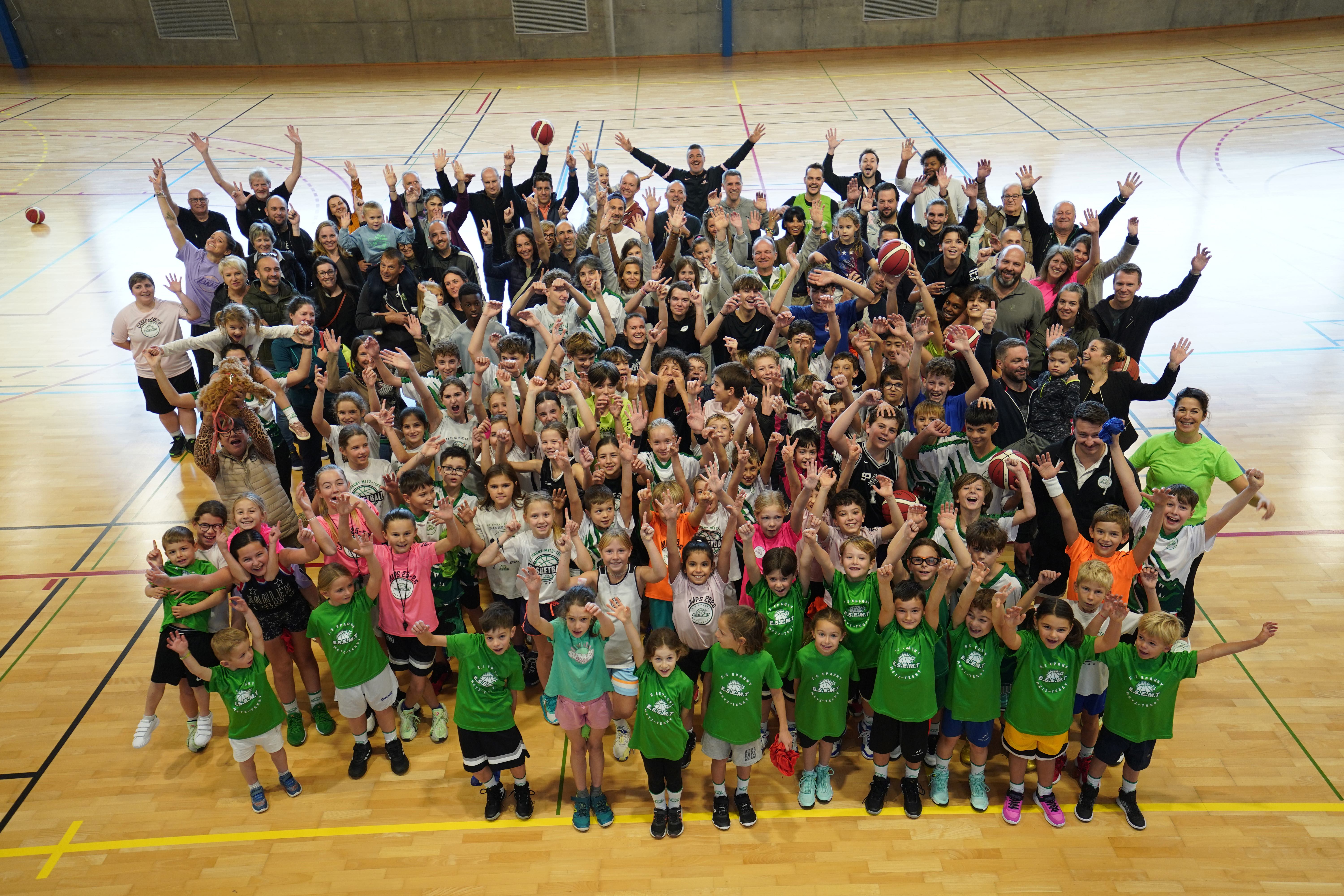 Photo de groupe vue d’en haut : jeunes licenciés en t-shirts verts de l’ESEMT, parents et coachs réunis sur le parquet, bras levés.